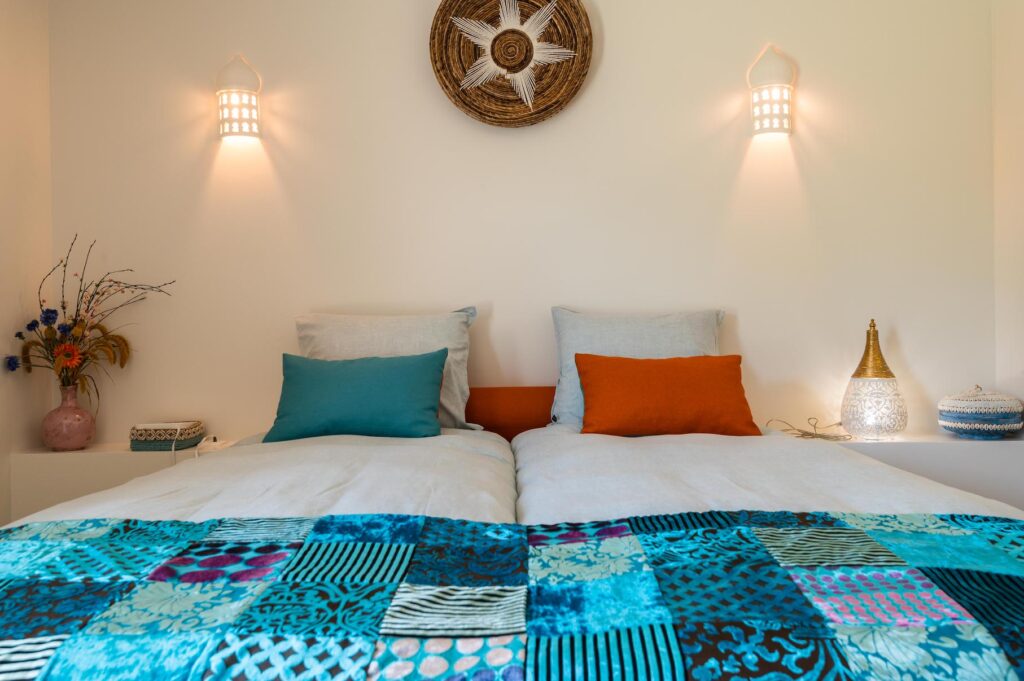 Gallery, Twin beds with colorful teal and orange pillows beneath a decorative wall mandala at the Seeds of Silence boutique hotel in Algarve, Portugal.