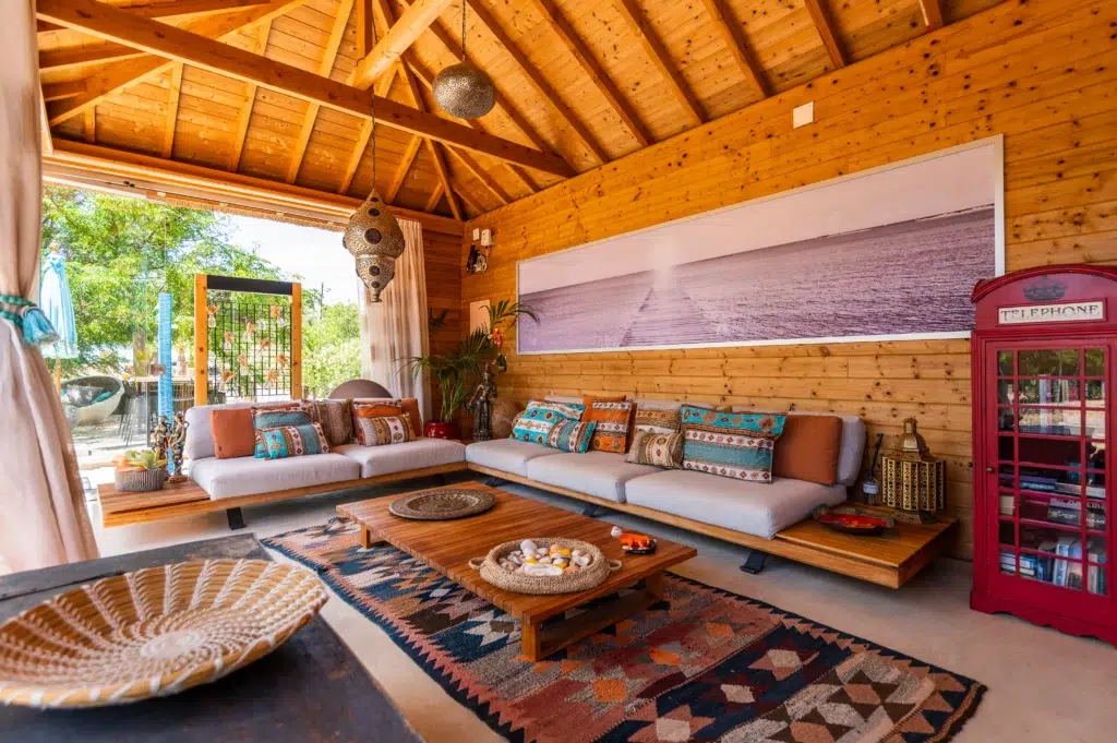 Spacious bohemian indoor lounge at Seeds of Silence featuring exposed wooden beams, plush low sofas with colorful pillows, a large panoramic wall photo, and a vintage red telephone booth.