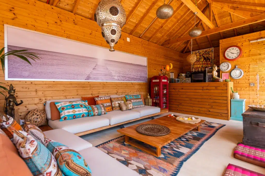 Sunlit bohemian indoor lounge at Seeds of Silence with wooden paneled walls, cathedral ceiling, plush low sofa with colorful cushions, and a large beach