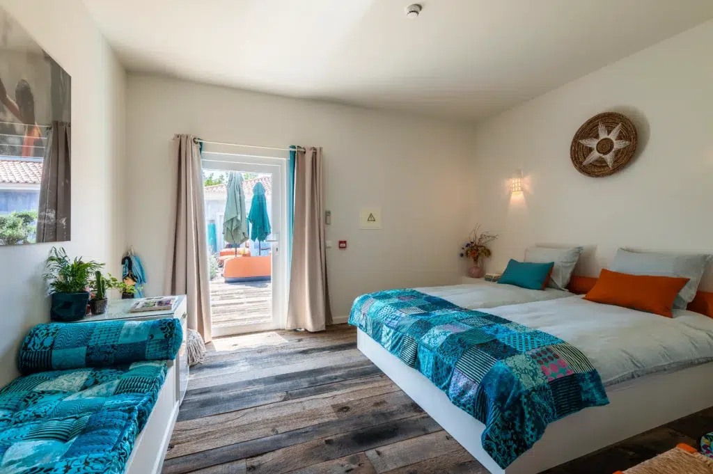 Bright boutique hotel guest room at Seeds of Silence featuring a bohemian blue patchwork bedspread, wooden flooring, and a glass door opening to an outdoor patio with umbrellas.