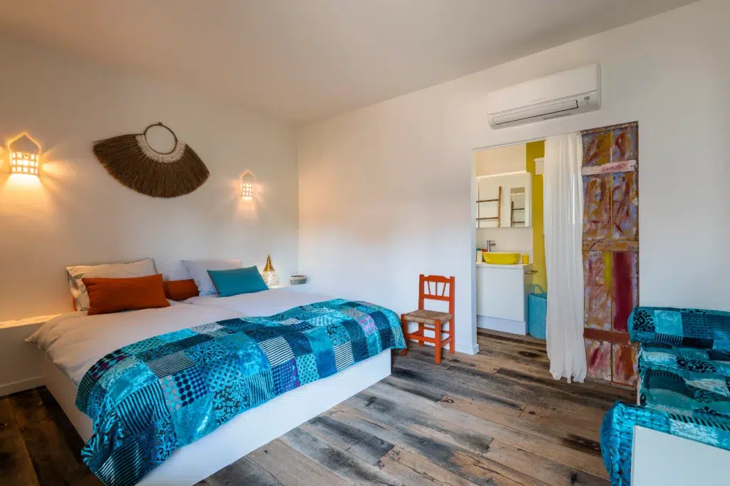 Bright boutique hotel bedroom at Seeds of Silence featuring a bohemian blue patch-work bedspread, wooden flooring, rustic sliding door, and attached modern bathroom.