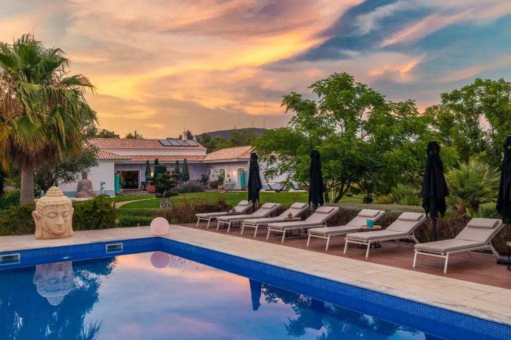 Luxury outdoor swimming pool at Seeds of Silence boutique hotel at sunset, with rows of lounge chairs, a Buddha head sculpture, and a vibrant orange sky.