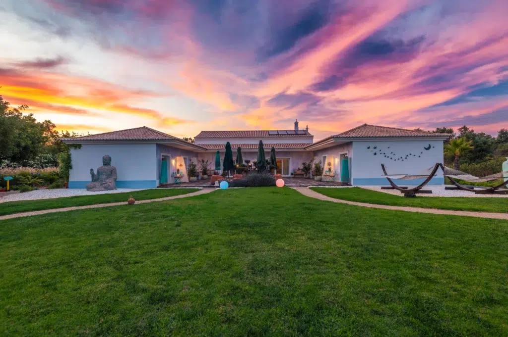 Wide sunset view of the boutique resort building at Seeds of Silence with a green lawn, hammock, and a stone Buddha sculpture by the pool.