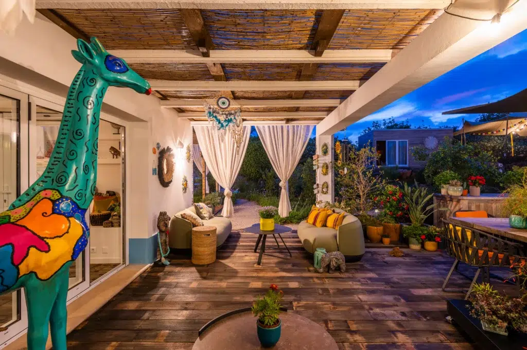 Evening view of a cozy covered outdoor patio at Seeds of Silence boutique hotel with wooden decking, plush seating, bohemian decor, a colorful giraffe sculpture, and a lit garden.