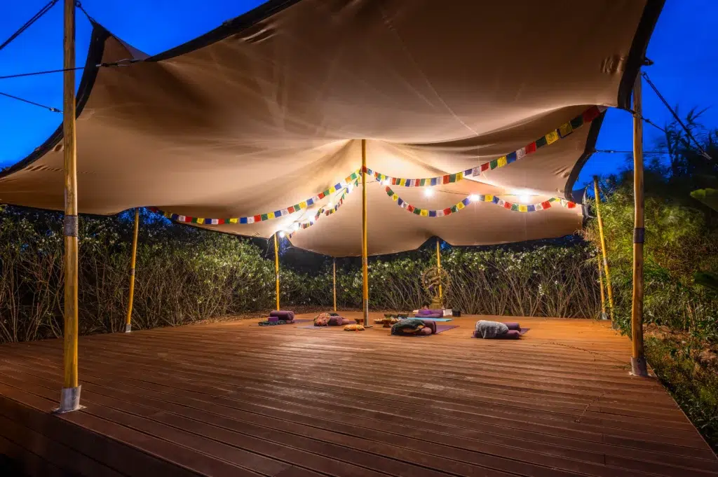 Covered outdoor yoga shala and meditation deck at Seeds of Silence boutique hotel at dusk, featuring yoga mats, bolsters, and prayer flags under a stretch tent.
